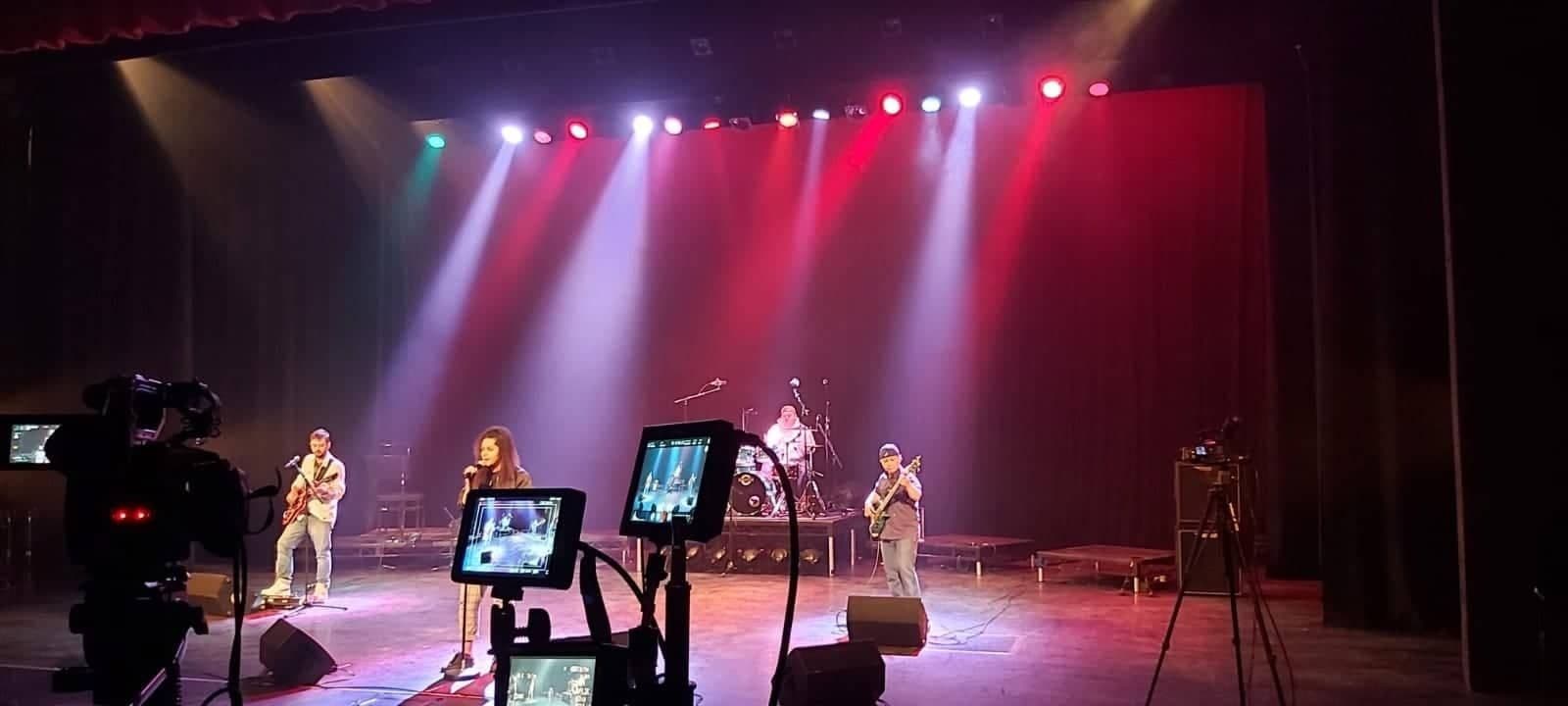 Band on a theatre stage under red and white stage lighting with cameras filming
